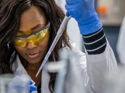 Student wearing lab coat and safety goggles injecting liquid into small container at the College of Science and Mathematics